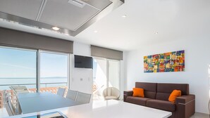 A modern living room with a large island, a sofa with orange cushions, a colorful abstract painting, and a large window overlooking the sea. - Modern Apartment with Panoramic Sea View & Terrace, by the Beach – Costa Brava (Roses)