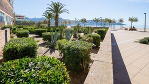 A landscaped outdoor area with a paved walkway, palm trees, and a view of the sea and mountains in the background. - Modern Apartment with Panoramic Sea View & Terrace, by the Beach – Costa Brava (Roses)