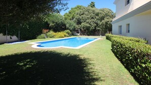 Une piscine entourée d’une pelouse et de haies bien entretenues, avec un bâtiment blanc en arrière-plan.