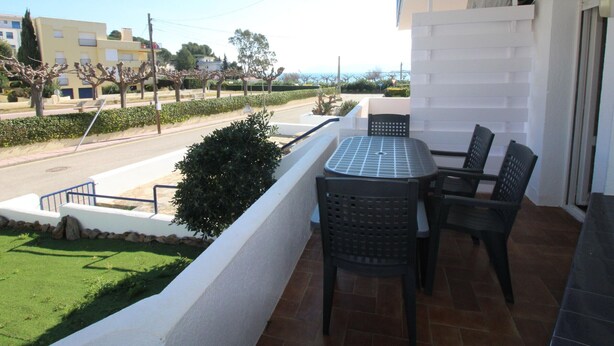 Uma varanda com mesa e cadeiras, vista para a rua e edifícios, e um espaço com grama.