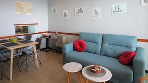 A living room with a blue sofa, two red cushions, a small table with a basket, and a dining area with a table and chairs. - Apartment with Terrace 300m from Beach, Parking Included (Llançà)