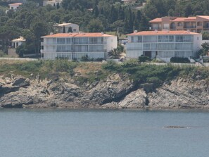 Un edificio frente al mar con techo de tejas rojas, amplias ventanas y un litoral rocoso en primer plano.