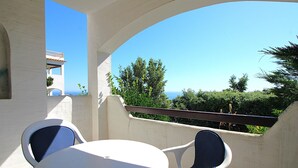 Un balcon avec une table blanche et deux chaises, d’où l’on peut admirer un paysage pittoresque avec des arbres et un bâtiment au loin.