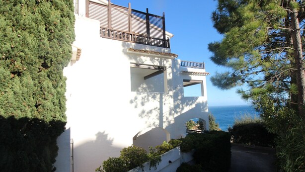 Un bâtiment blanc doté d’un balcon, entouré de verdure et donnant sur une étendue d’eau.