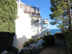 Un bâtiment blanc doté d’un balcon, entouré de verdure et donnant sur une étendue d’eau.