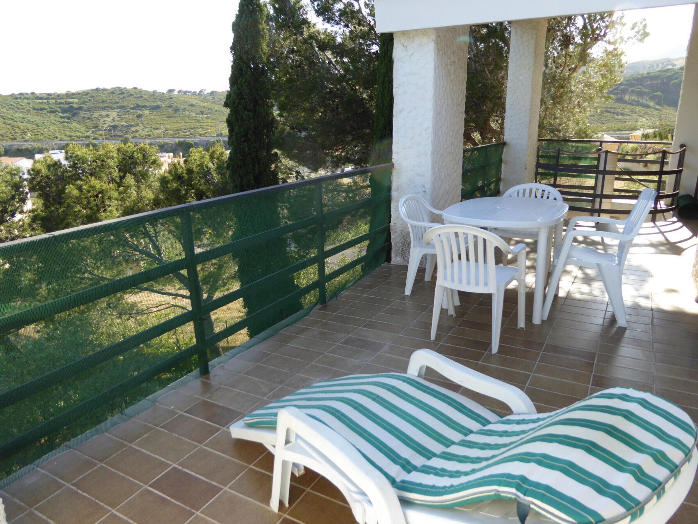 A balcony with a table and chairs, a striped lounge chair, and a view of a green landscape.