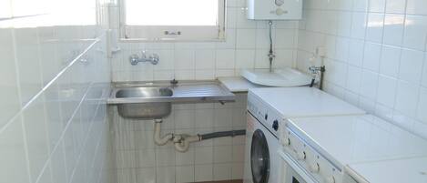 A compact kitchen with white tiles, a stainless steel sink, a washing machine, and a built-in oven.