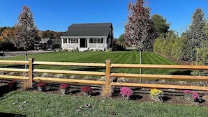 A house with a porch, surrounded by a wooden fence and a well-maintained lawn with colorful flowers. - Beach house, inground pool, hot tub, Large backyard,1.5 Miles from Misquamicut (Westerly)