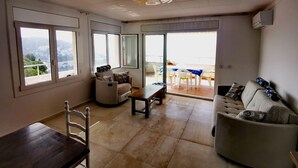 A spacious living room with a sofa, a dining table, and chairs. Large windows provide a view of the outdoors.