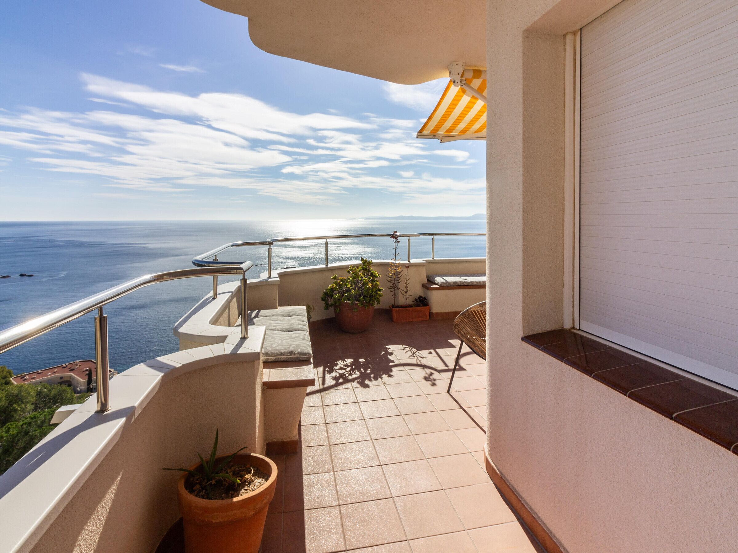 A balcony with a view of the sea, a potted plant, a cushioned seating area, and a closed sliding door.