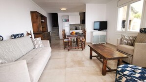 A living room with a sofa, a wooden coffee table, and a patterned ottoman. There is a dining area with a table and chairs, and a kitchen in the background.