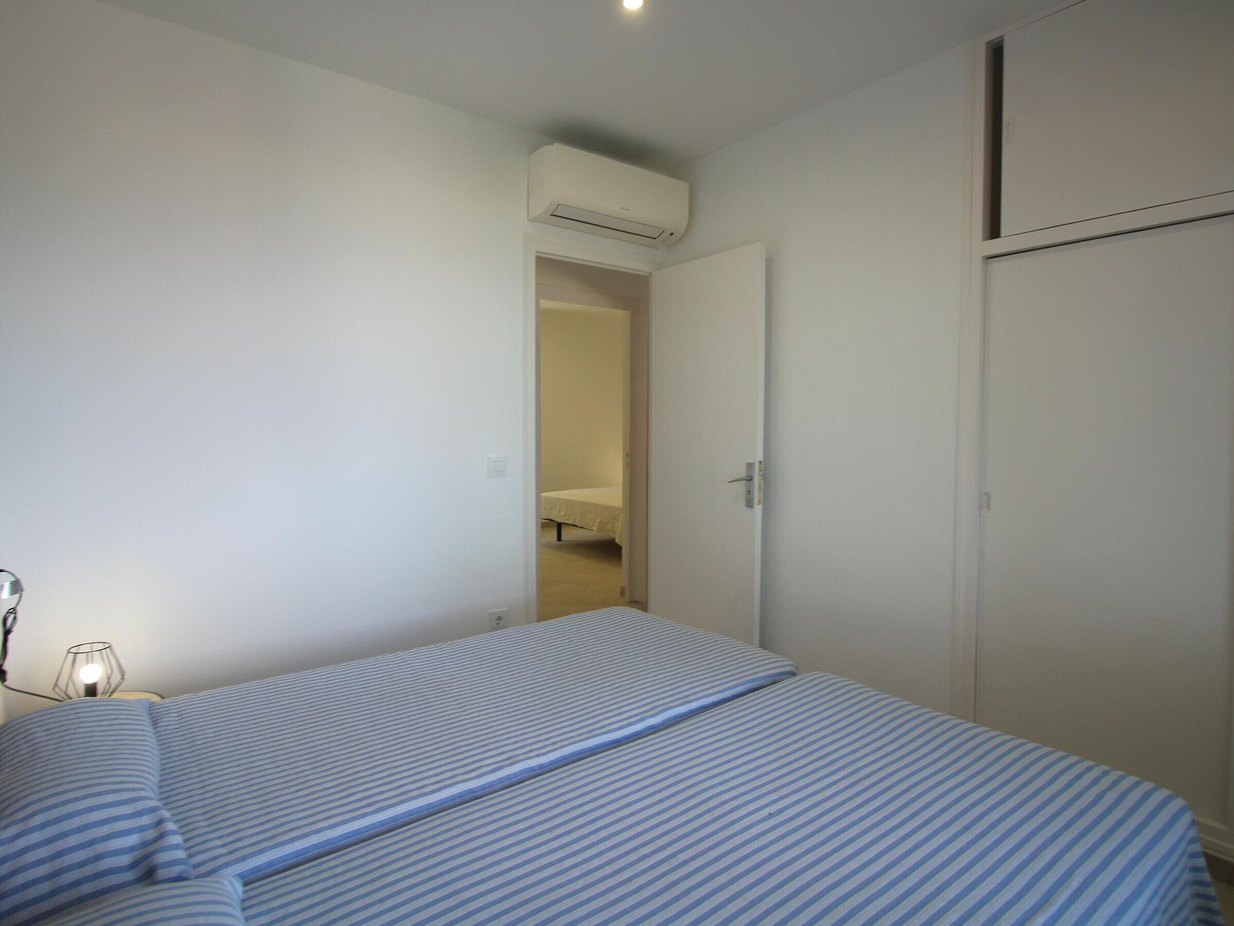 A bedroom with a bed, a bedside table, an air conditioning unit, and a door leading to another room.
