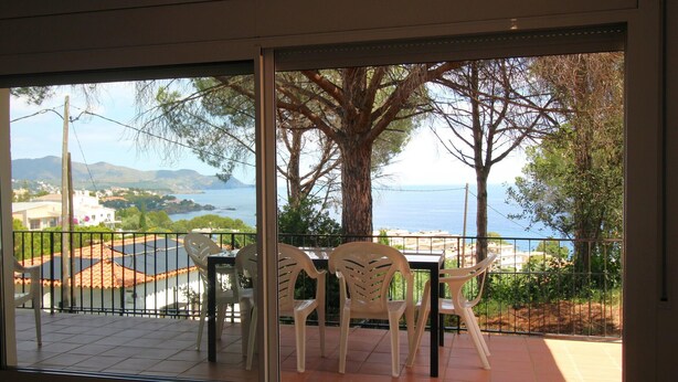 Varanda com duas cadeiras brancas e uma mesa, com vista para uma área costeira arborizada e o mar.