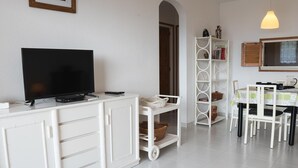 A living room with a television on a white cabinet, a white bookshelf, a dining table with chairs, and a hanging light fixture. - Bright apartment with pool and terrace, near the beach (Llançà)