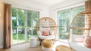 A bright room with large windows, a wicker chair, a white sofa, and a round ottoman. - Sunroom, Spacious Deck, 8 Mins to Mayflower Beach! (Dennis)