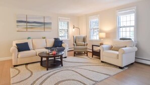 A living room with a beige sofa, a round coffee table, and two armchairs. There are large windows with white frames and a ceiling vent. - Sunroom, Spacious Deck, 8 Mins to Mayflower Beach! (Dennis)
