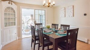 A dining area with a wooden table, dark chairs, and a chandelier. There is a white cabinet with glass doors and a view of the outdoors through a glass door. - Sunroom, Spacious Deck, 8 Mins to Mayflower Beach! (Dennis)