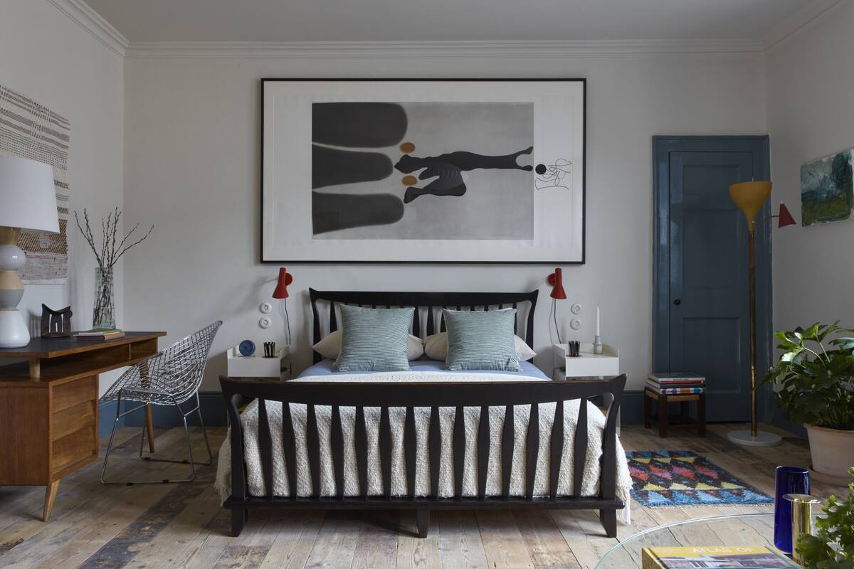 Un dormitorio con cama, escritorio, silla, lámpara, una obra de arte enmarco y una puerta.