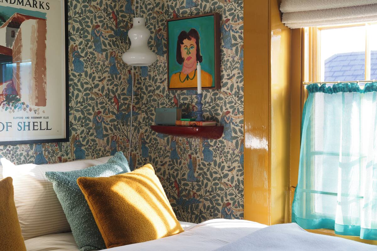 Un dormitorio con papel tapiz floral, un cuadro enmarco, una lámpara de noche y una ventana con cortinas.