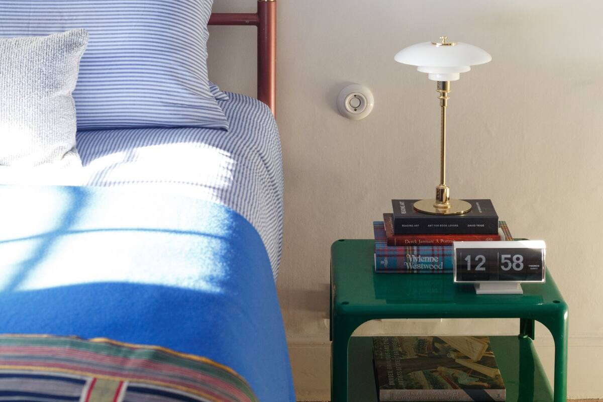 Una cama bien tendida con ropa de cama azul, una mesita de noche verde con una lámpara y libros, y un cuadro abstracto enmarcado en la pared.