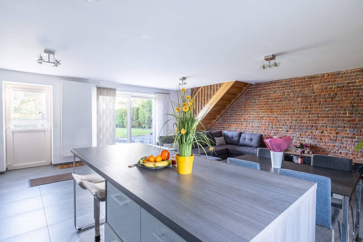 A modern kitchen with a large island, a brick wall, and a staircase leading to a loft.