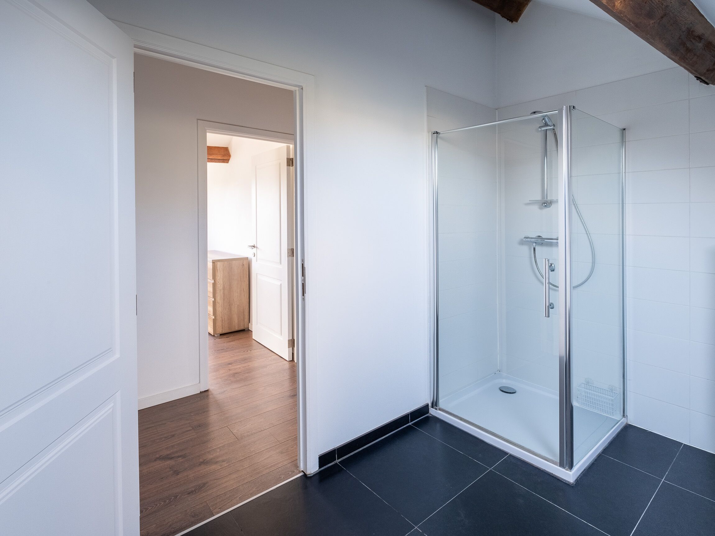 A modern bathroom with a glass shower enclosure, dark tiled flooring, and a wooden cabinet in the adjacent room.