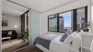 A modern bedroom with a large window, a bed, a sofa, and a view of a cityscape.