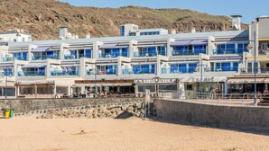 Un edificio costero con varios balcones, una playa arenosa al frente y una ladera rocosa en el fondo.