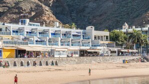 Un hotel frente al mar con varios balcones, una playa de arena y una ladera rocosa al fondo.