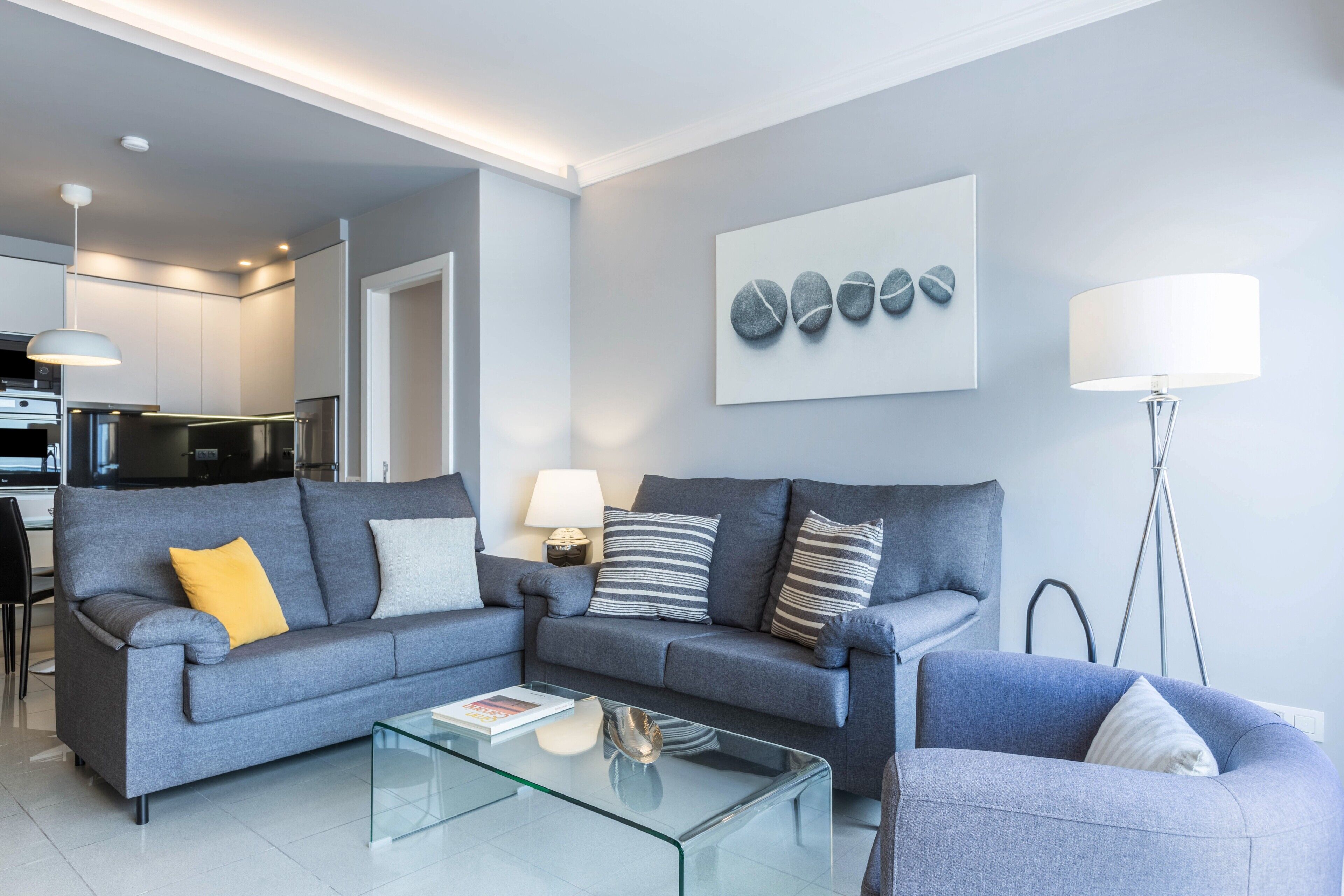 Una sala de estar moderna con un sofá gris, una mesa de centro de vidrio y una cocina al fondo.