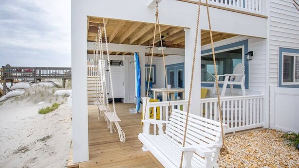 Une maison de plage dotée d’une terrasse en bois, de chaises suspendues blanches et d ’un porche abrité.