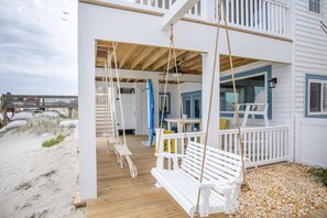Una casa de playa con terraza de madera, sillones blancos columpios y un porche cubierto.