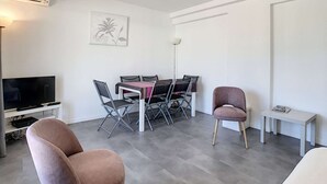 A modern living room with a flat-screen TV, a dining table with chairs, a sofa, and a small table with a lamp. - Berlioz Garden - Jardin,Free Parking,AC,Wifi (Antibes)