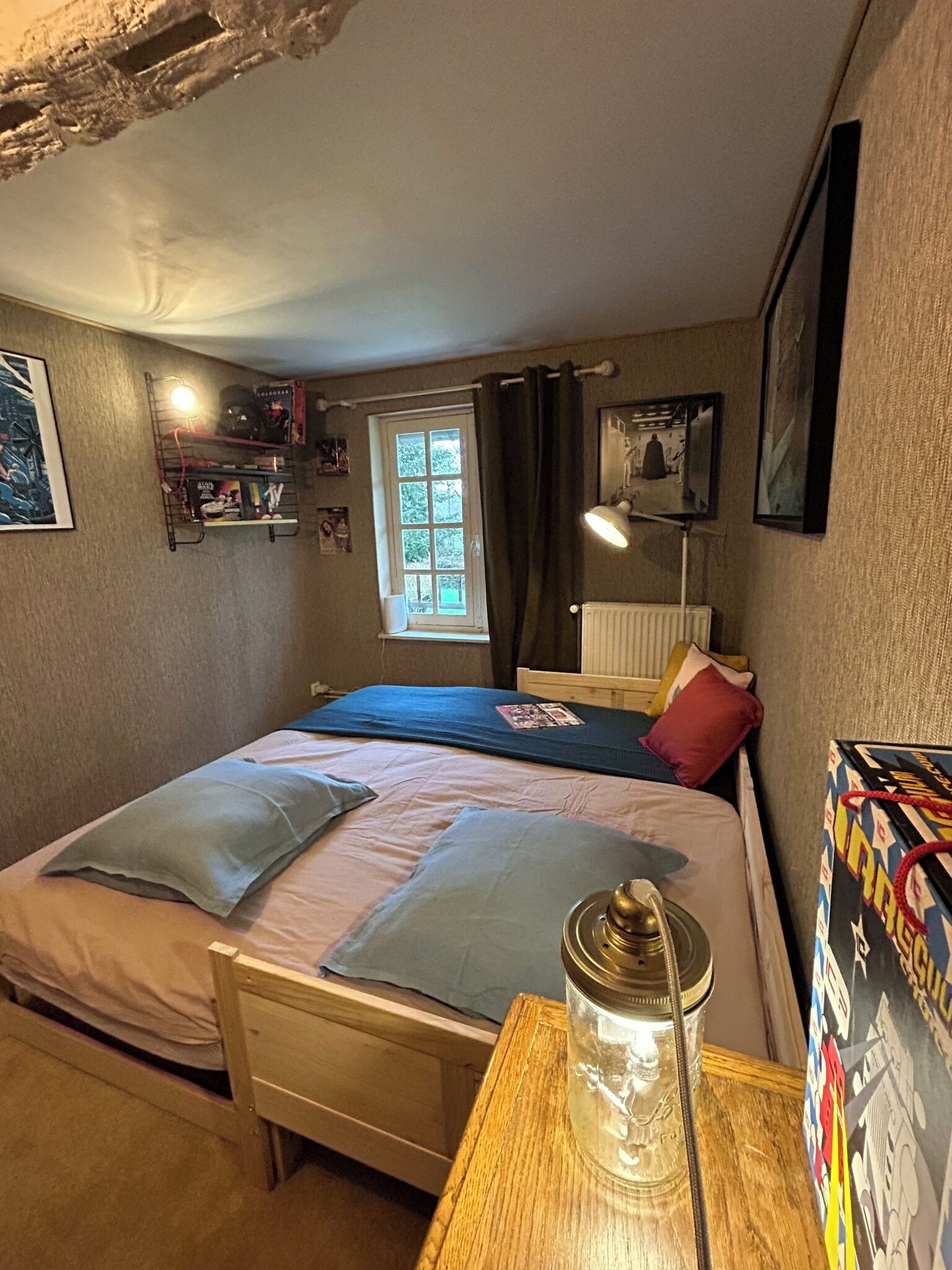 A small, cozy bedroom with a single bed, a nightstand, a lamp, and a window with curtains.