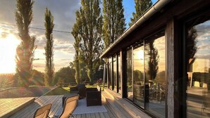 A wooden deck with outdoor furniture, large windows, and a view of trees and the sky. - Domaine de Blanvatel (Saint-Pierre-en-Auge)