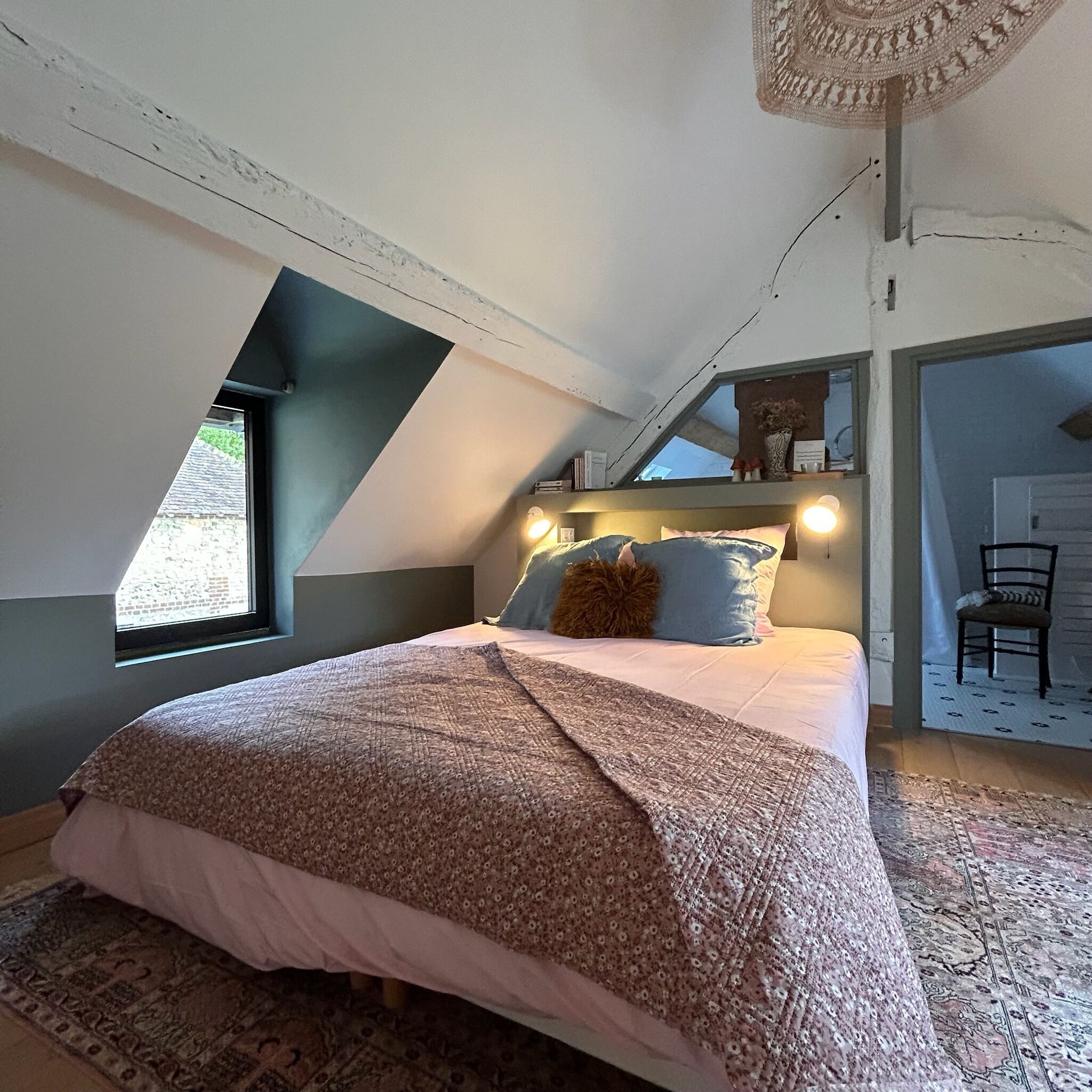 Un dormitorio con techo inclinado, una cama con colcha estampada, una ventana y una silla en la esquina.