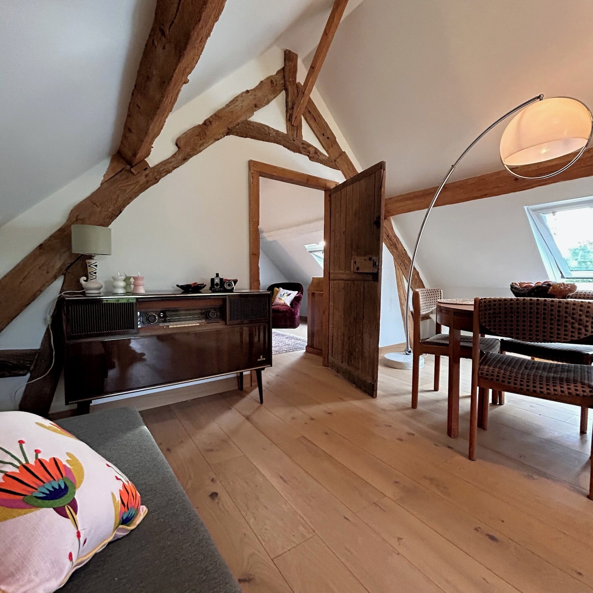 Ein gemütliches Dachzimmer mit freiliegenden Holzbalken, einem Esstisch mit Stühlen, einem Vintage-Radio und einem Fenster mit Blick.