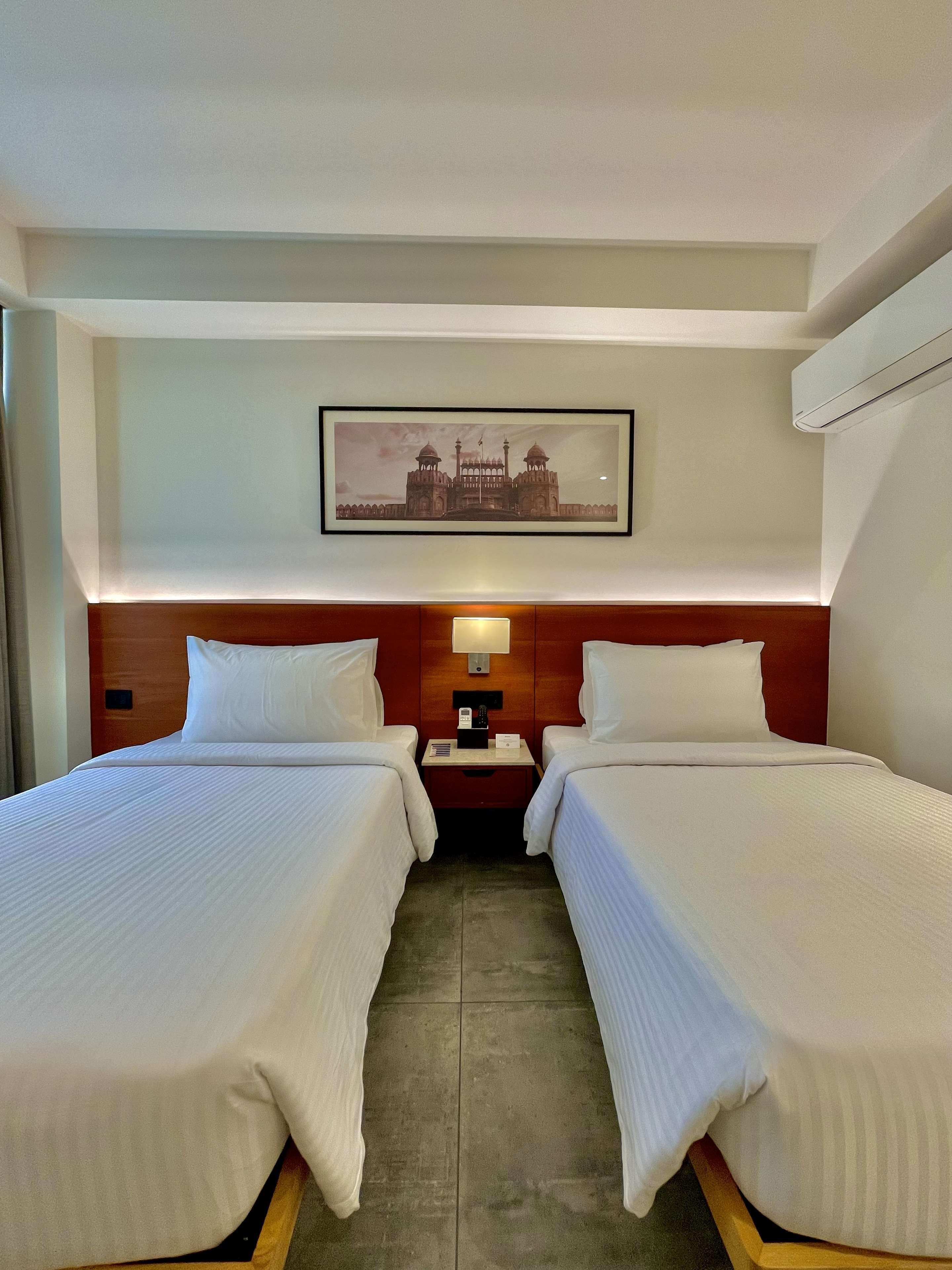 A hotel room with two beds, a nightstand, a wall-mounted lamp, and a framed picture of a historical building.