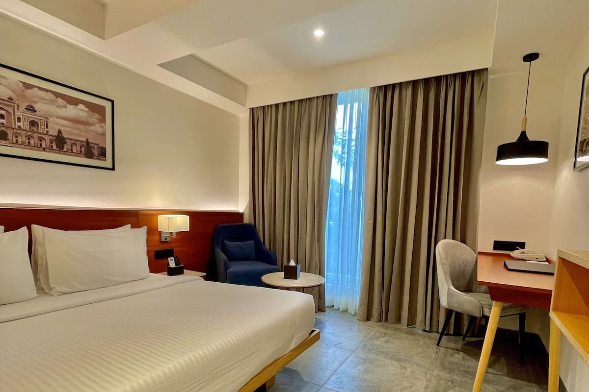 A hotel room with a large bed, a desk, two chairs, a small table, a lamp, and a painting on the wall.