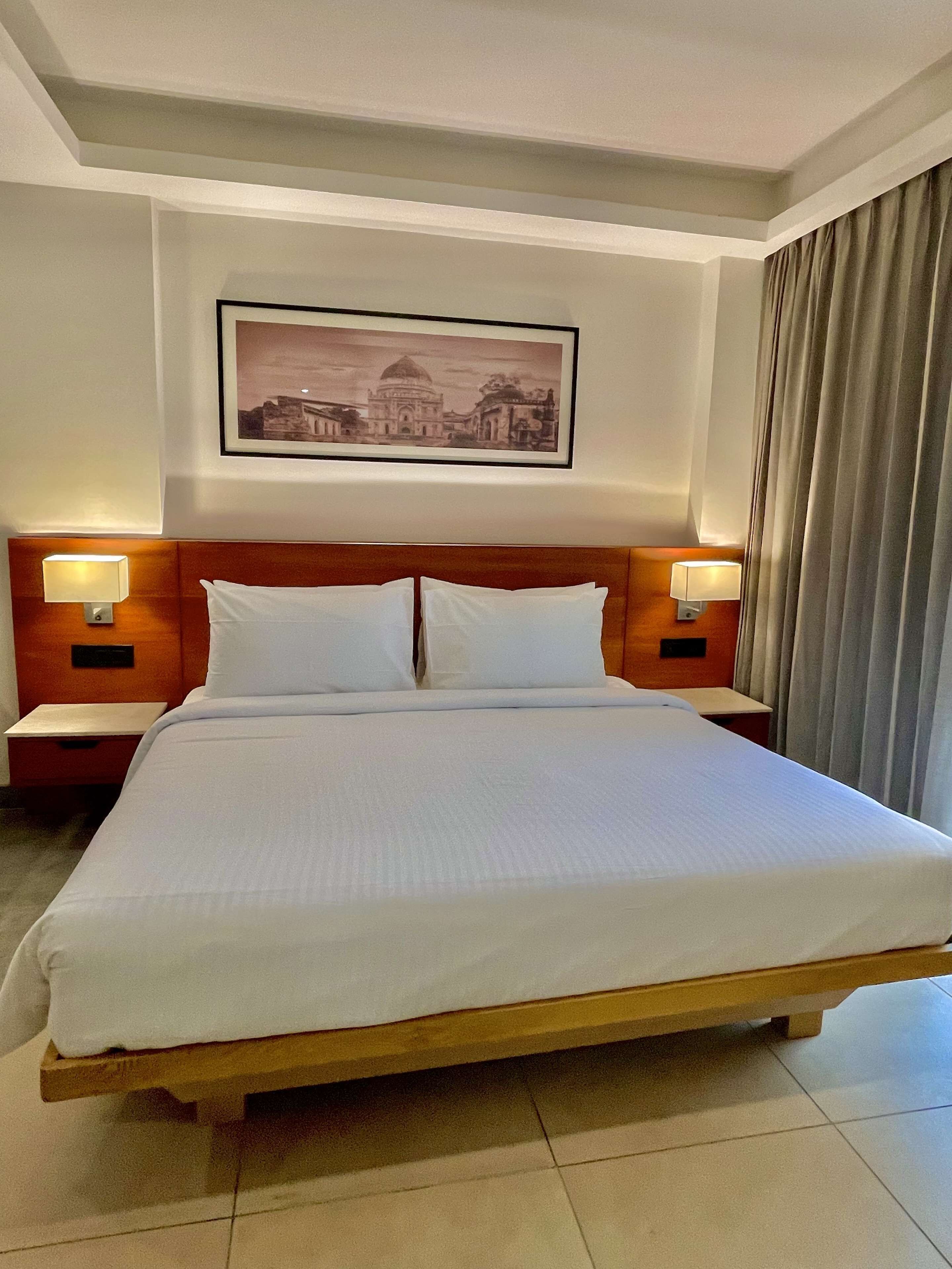 Ein Hotelzimmer mit einem großen Bett, zwei Nachttischen mit Lampen, einem gerahmten Bild an der Wand und einem Vorhang am Fenster.