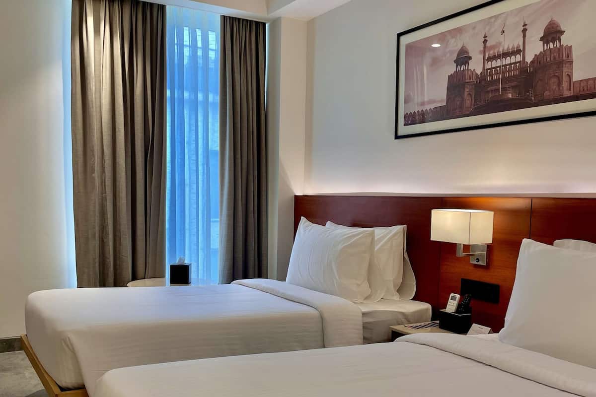 A hotel room with two beds, a nightstand, a lamp, a framed picture of a building, and a window with curtains.