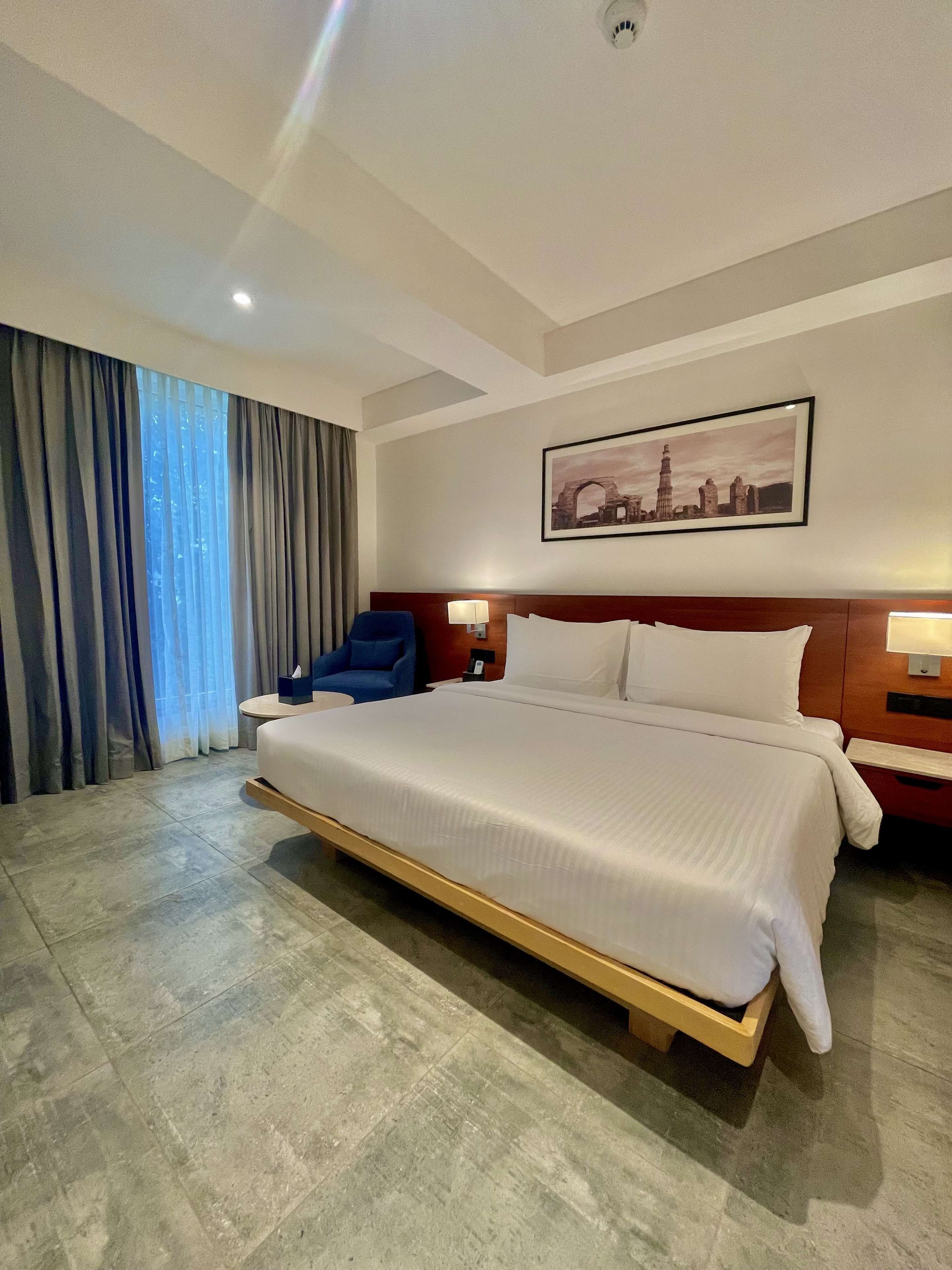 Amplia habitación de hotel con una cama grande, un sillón azul, una mesita de noche y un cuadro de un paisaje urbano en la pared.