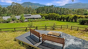 A wooden deck with two benches and a table, overlooking a grassy field and a building with a sloped roof. - Kookaburra Hill, Kangaroo Valley (Kangaroo Valley)