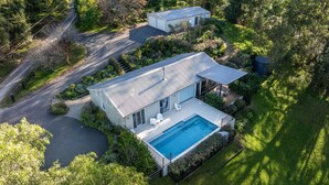 Une maison moderne à deux étages, dotée d’une piscine, entourée d’une végétation luxuriante et accessible par une allée pavée.