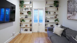 Een woonkamer met een grijze bank, houten vloeren, witte wandplanken met boeken en planten, een aan de muur bevestigde televisie en een deur met ramen.