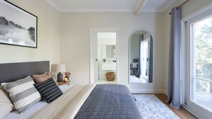 Een slaapkamer met een groot bed, een nachtkastje met een lamp, een lijst met een afbeelding aan de muur, en zicht op de badkamer door een boogvormige deuropening.