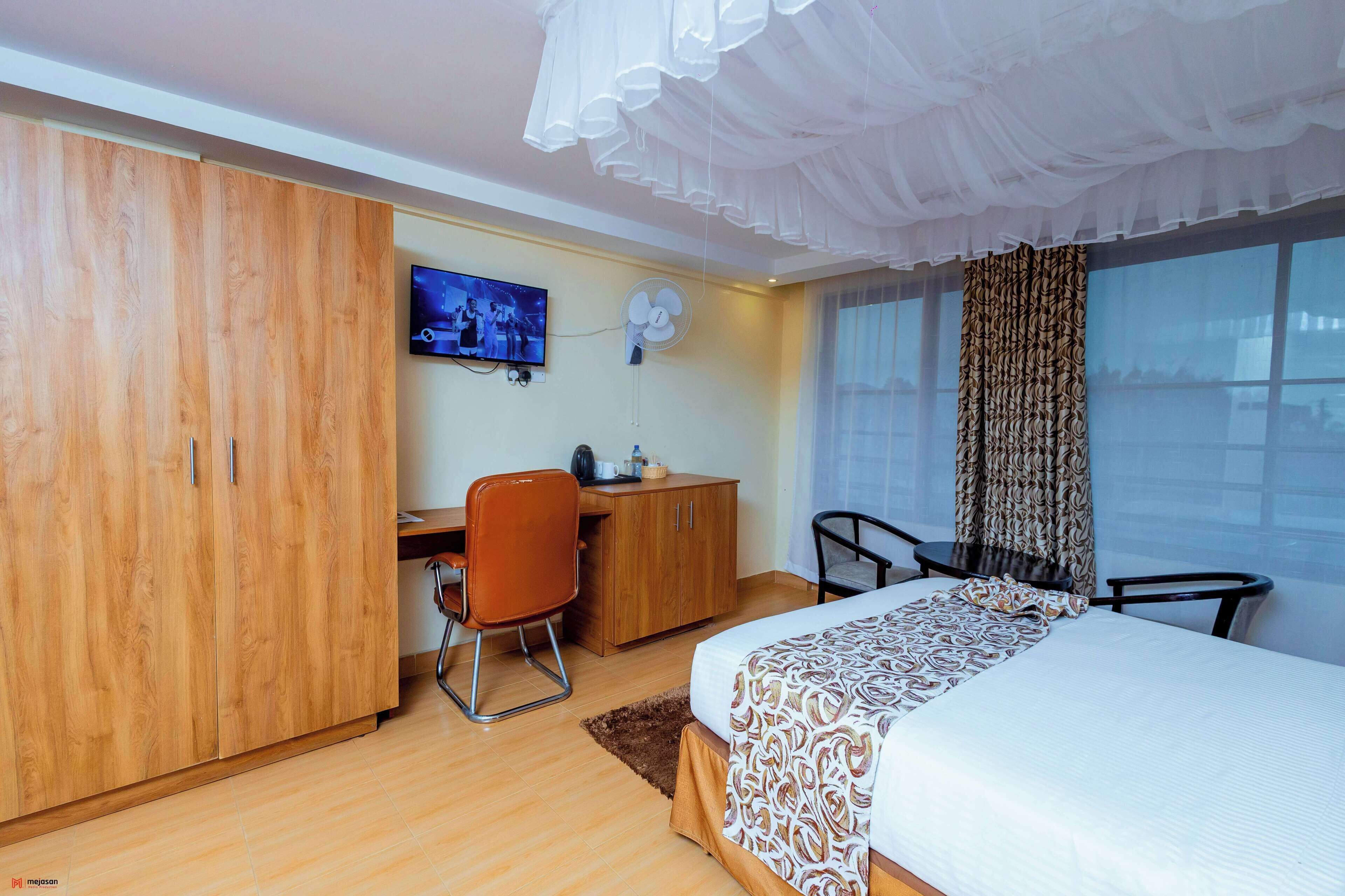 Quarto de hotel com cama, escrivaninha, cadeira, guarda-roupa, televisão e janela com cortinas.