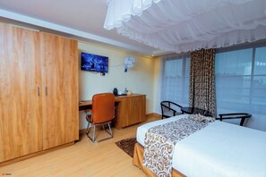 Habitación de hotel con cama, escritorio, silla, armario, televisión y ventana con cortinas.