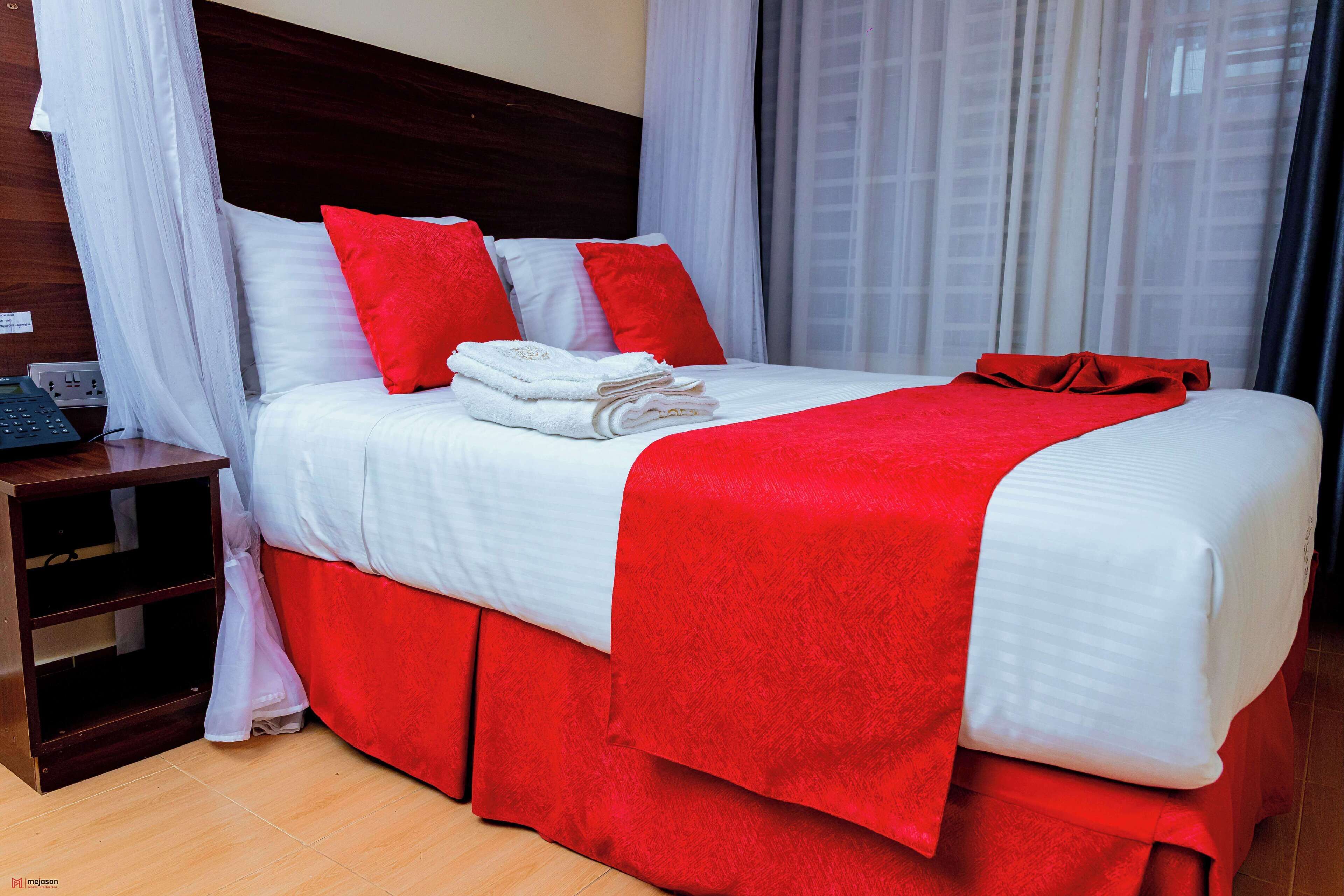 Ein Hotelzimmer mit Bett, weißer Bettwäsche, roten Akzenten, einem Holzkopfschild, einem Nachttisch und Blick auf die Stadt durch leichte Vorhänge.