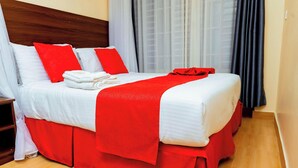 A hotel room with a neatly made bed, a wooden headboard, and a red accent on the bedspread.